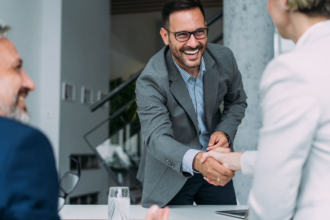 A smiling man in a gray blazer and glasses shakes hands with a woman in a white suit jacket, discussing B2B marketing KPIs during a meeting. Another man in a blue suit sits to the side, also smiling. They are in a modern office with a staircase in the background.