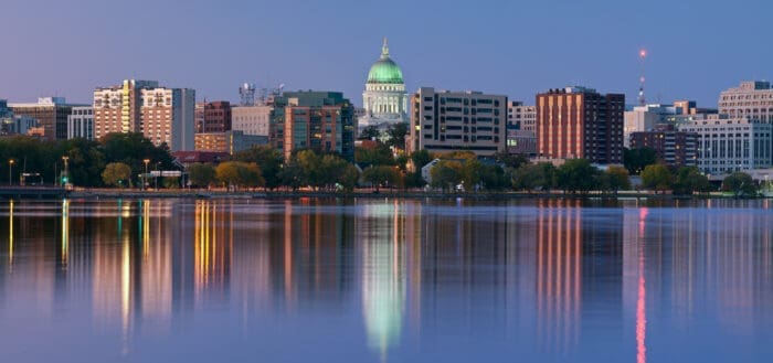 A serene evening view of the Wisconsin city skyline features a prominent capitol building with a green dome, surrounded by various buildings. The reflection of the city lights shimmers on the calm water in the foreground.