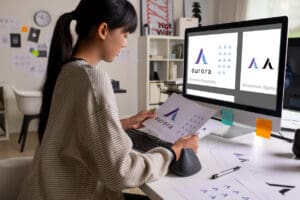 A woman sits at a desk, meticulously working on the logo design for a brand called "Aurora." She holds a printed logo sheet and uses a computer with the same logo on the screen. The workspace is clean and modern, featuring various design elements and notes that adhere to top-notch logo design principles.