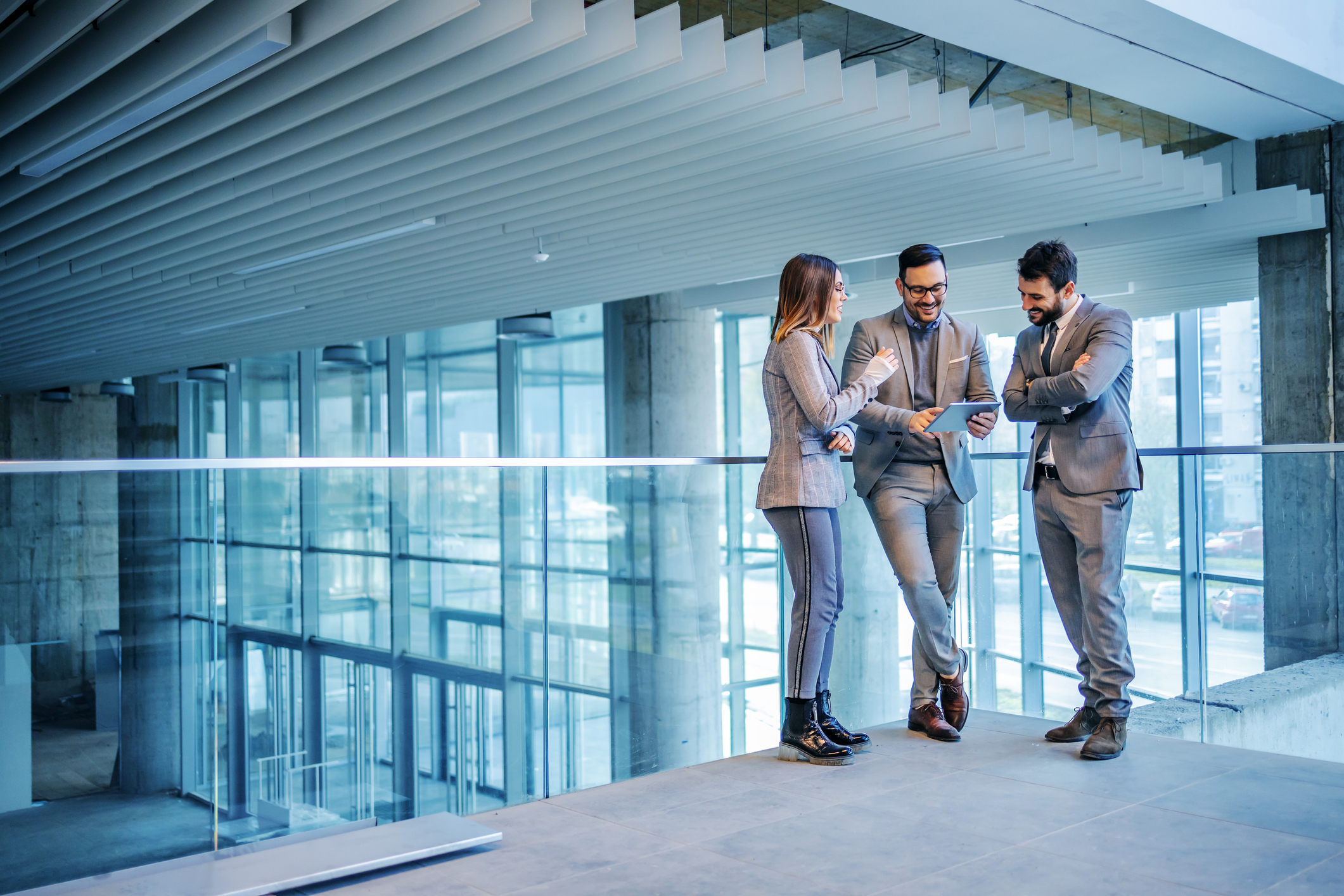 Three people in business attire stand in a modern office building. One is holding a tablet, and they all appear to be engaged in discussing marketing agency strategies. Glass walls and a contemporary ceiling structure are visible in the background.