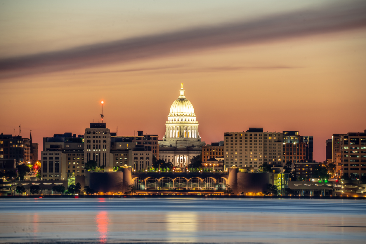 A distant view of a city skyline at sunset, featuring a prominent white-domed building in the center. The sky is a gradient of warm colors, beautifully illustrating why Madison's website design is inspired by nature. The blurred water surface reflects the vibrant city lights, creating an enchanting scene.