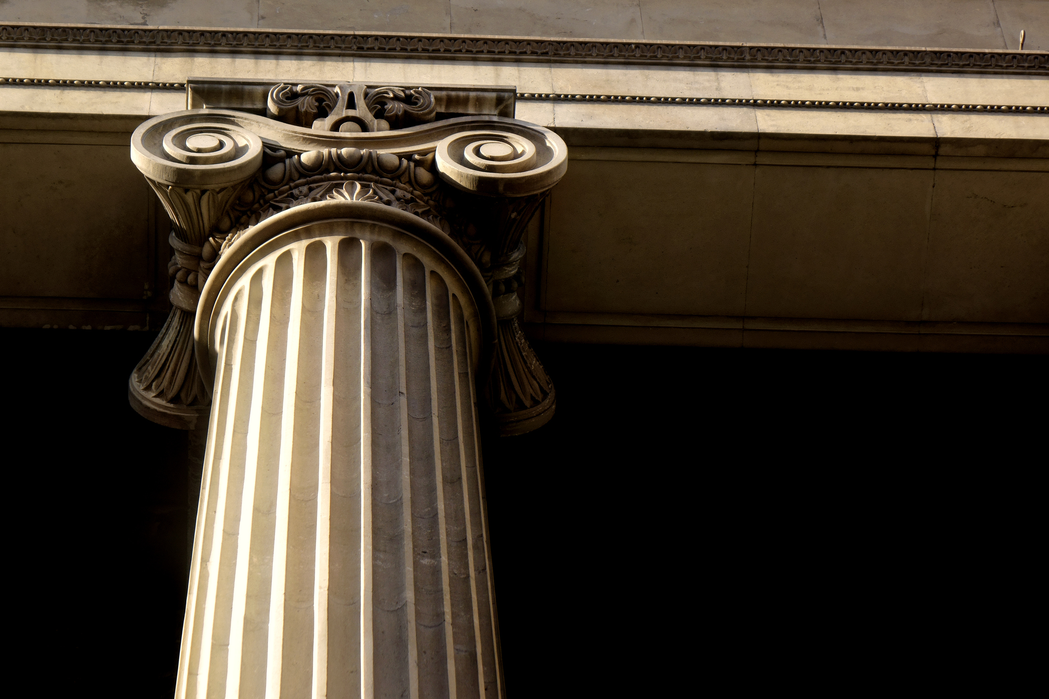 A close-up of a Corinthian column with intricate carvings and decorative scrolls, resembling the structure of a pillar page. The image highlights the column’s detailed capital and fluted shaft set against a dark background, capturing classical architectural design.