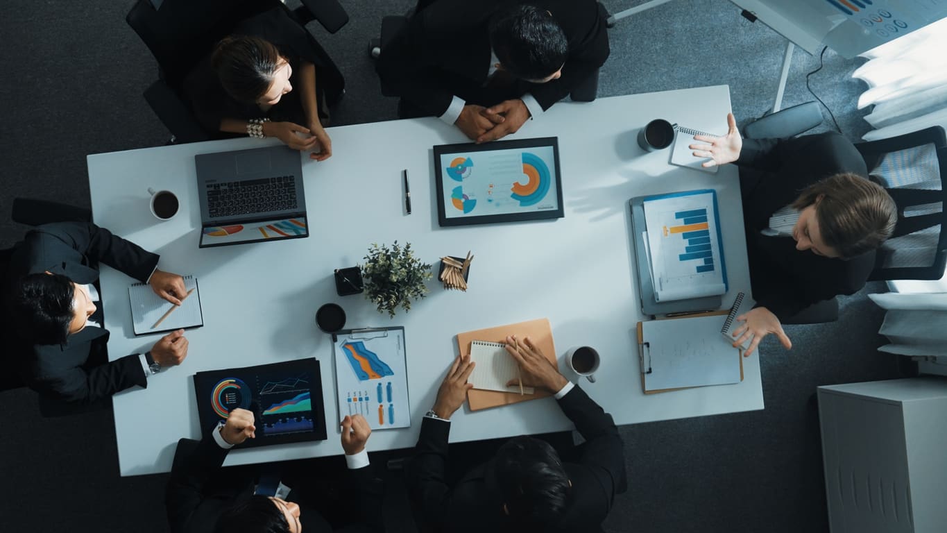 A group of people in business attire sits around a rectangular table in a strategy planning meeting. Laptops and documents displaying charts and graphs are spread across the table. A potted plant and coffee cups are also visible, enhancing the collaborative atmosphere.