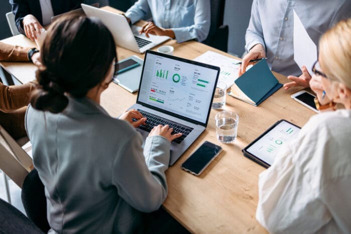 A group of people sit around a wooden table in a meeting room, working on laptops and tablets displaying search engine marketing charts and graphs, with documents, notebooks, and glasses of water also on the table.
