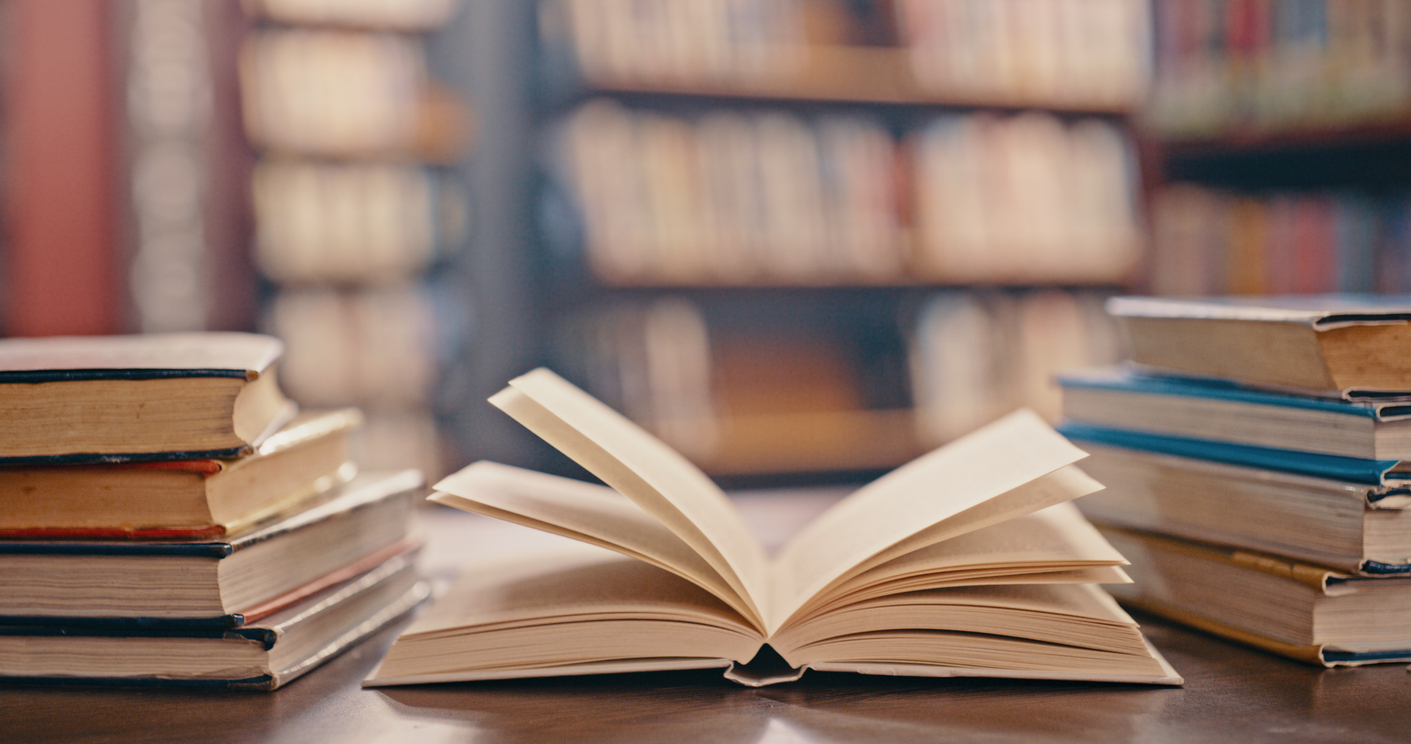 An open book on a wooden table is surrounded by stacks of closed business strategy books. Shelves filled with more volumes are blurred in the background, suggesting a library or study setting perfect for diving into strategic insights.