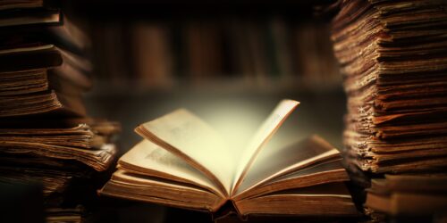 A dimly lit scene shows an open old book in the center, illuminated as if glowing. Surrounding it are tall stacks of weathered, aged books, alluding to the purpose of business and giving a sense of history and mystery. The background is blurred, making the book the focal point.