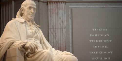 A marble statue of Benjamin Franklin seated and wearing historical attire is prominently displayed indoors. To the right, a wall features the inscription: "To err is human, to repent divine, to persist devilish." The setting is grand with ornate columns in the background, a testament to "Diligence is the Mother of Good Luck.